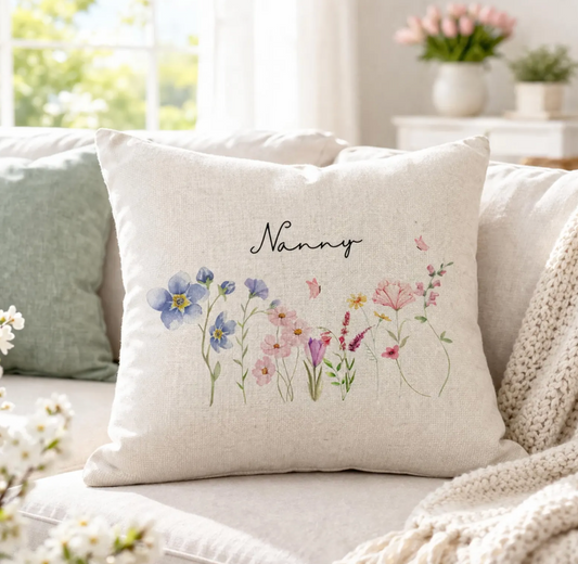 A square cream-colored throw pillow with a watercolor floral design and the word 'Nanny' in black script, resting on a light beige sofa in a bright, softly lit living room.