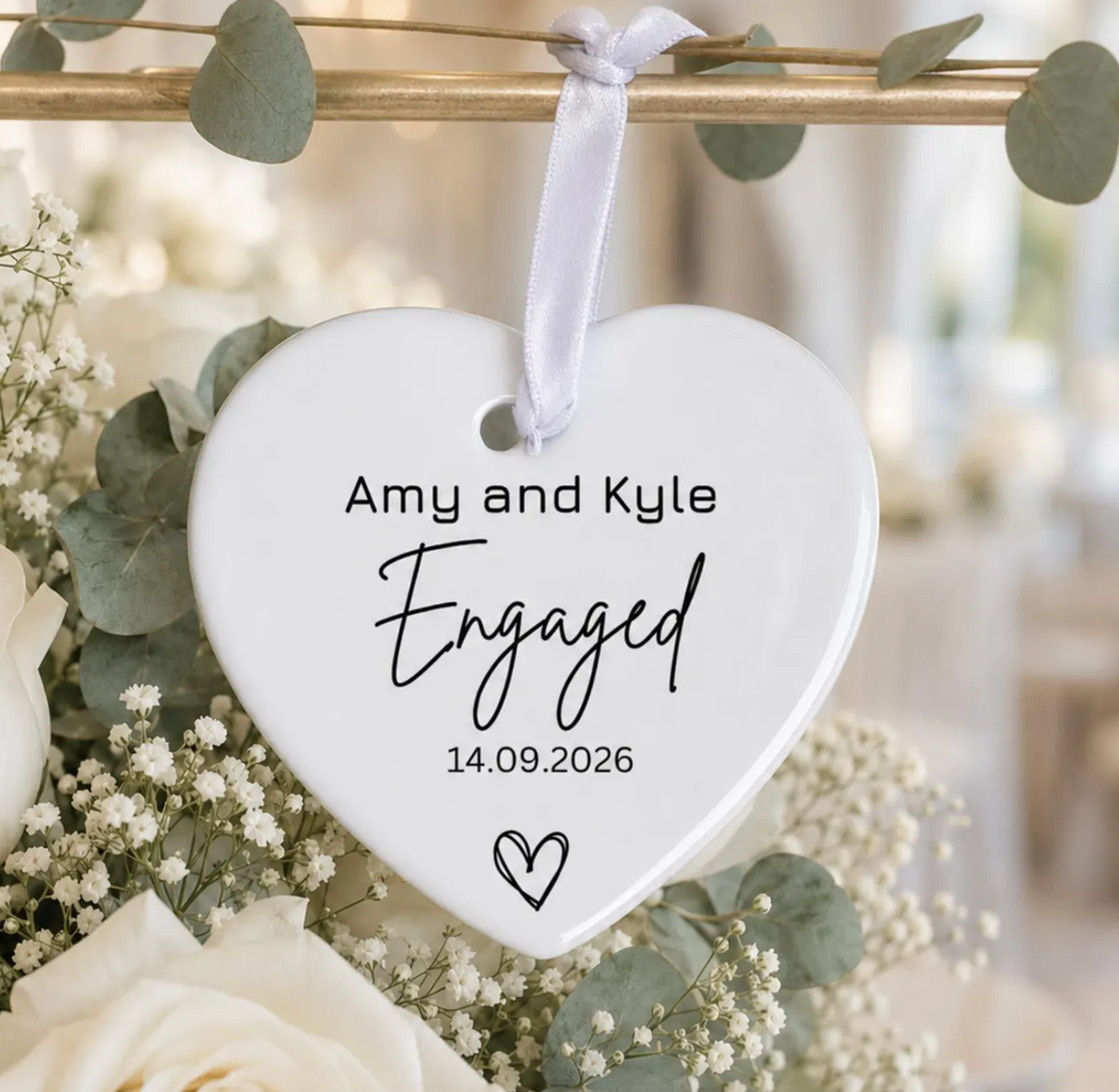 A white heart-shaped ceramic engagement plaque hanging from a white ribbon, decorated with black text reading 'Amy and Kyle Engaged 14.09.