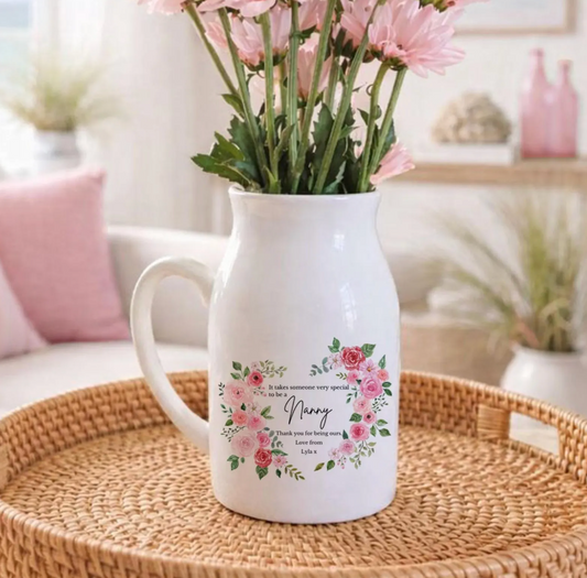 A white ceramic jug filled with fresh green tulip stems and buds, featuring a floral heart-shaped wreath design on the front. Inside the wreath, the text reads: "It takes someone very special to be a Nanny. Thank you for being ours. Love Jessie, Michael & Arthur xx." The jug is placed on a rustic wooden surface, surrounded by soft decor and greenery.