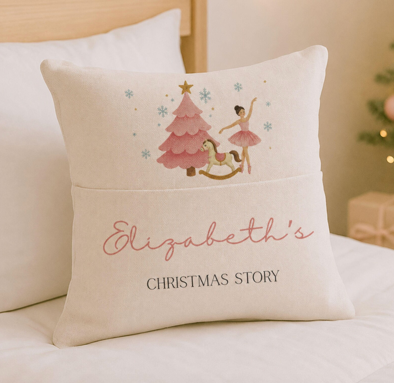 A personalized cream-colored throw pillow on a white bed, featuring a pink Christmas tree, ballerina, and rocking horse on the top half, with the text 'Elizabeth's CHRISTMAS STORY' on the bottom half.