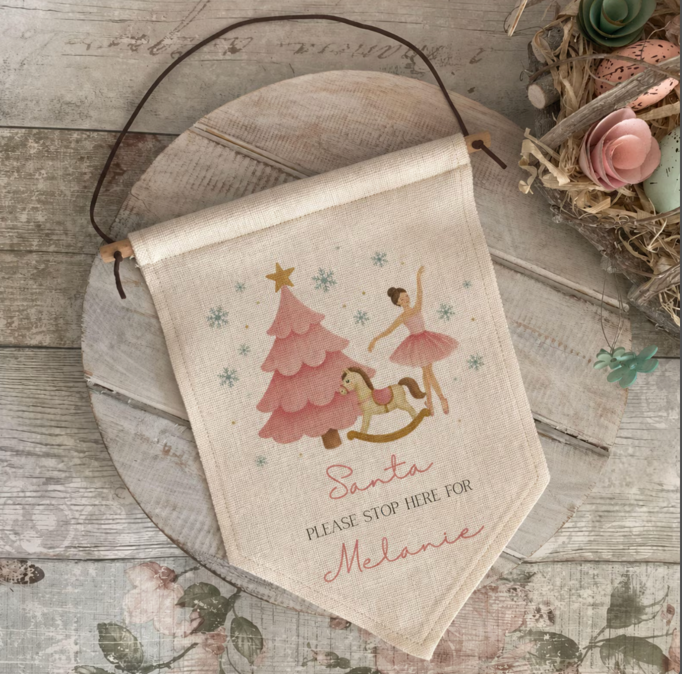 A personalized Christmas banner flag made of beige linen fabric, featuring a printed illustration of Santa, a ballerina, a rocking horse, and a Christmas tree, with the text 'Santa PLEASE STOP HERE FOR Melanie'. The flag is hung from a dark brown cord and displayed on a rustic wooden board with decorative dried flowers in the background.