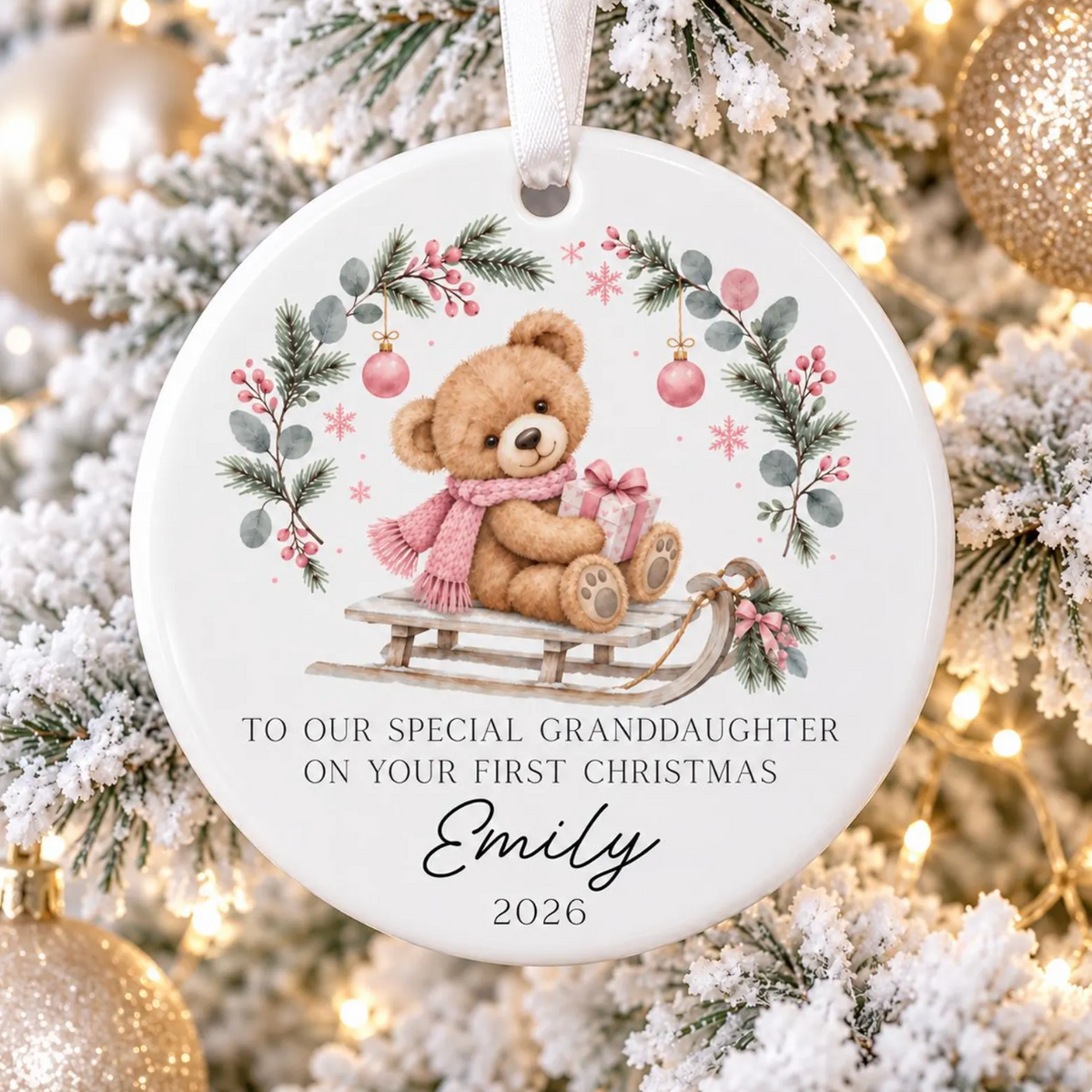 A round white Christmas ornament featuring a teddy bear sitting on a sled, surrounded by a floral wreath. The ornament has a personalized message for a granddaughter and is shown hanging against a decorated Christmas tree with lights and gold ornaments.