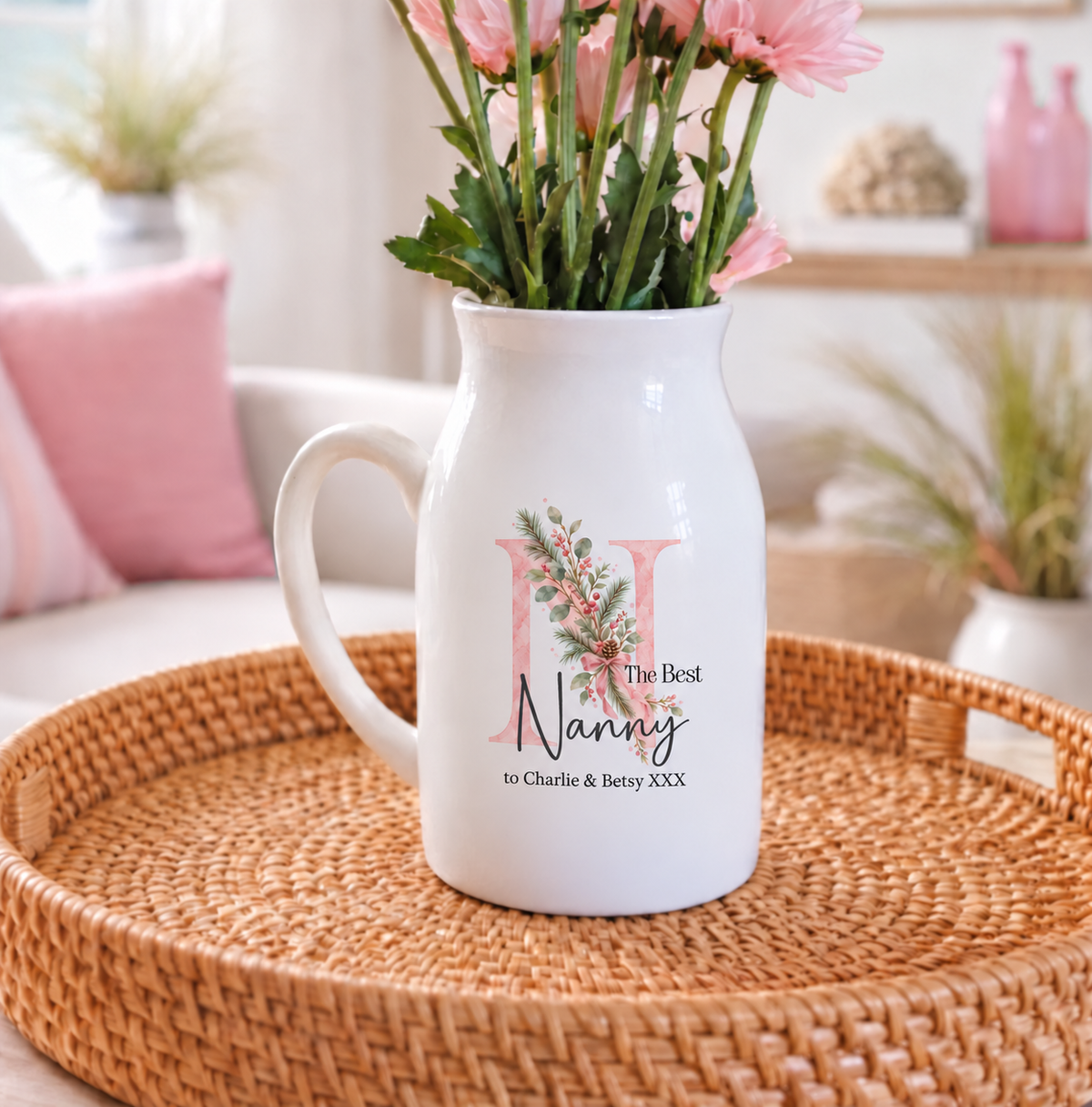 A white ceramic pitcher vase filled with pink flowers, placed on a woven round tray in a softly styled room. The front of the vase has a pink floral monogram and the words 'The Best Nanny to Charlie & Betsy XXX'.