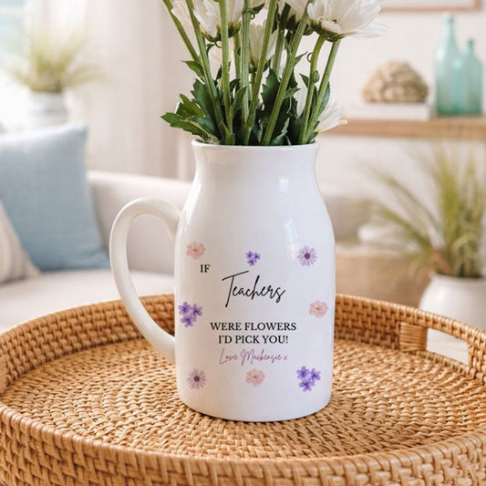 A white ceramic jug used as a vase, The jug is decorated with small purple and pink flowers and a printed message that reads: "If Teachers were flowers I’d pick you! Love Shauna xx." The jug is placed on a rustic wooden surface, with floral decorations and wooden barrels in the background.