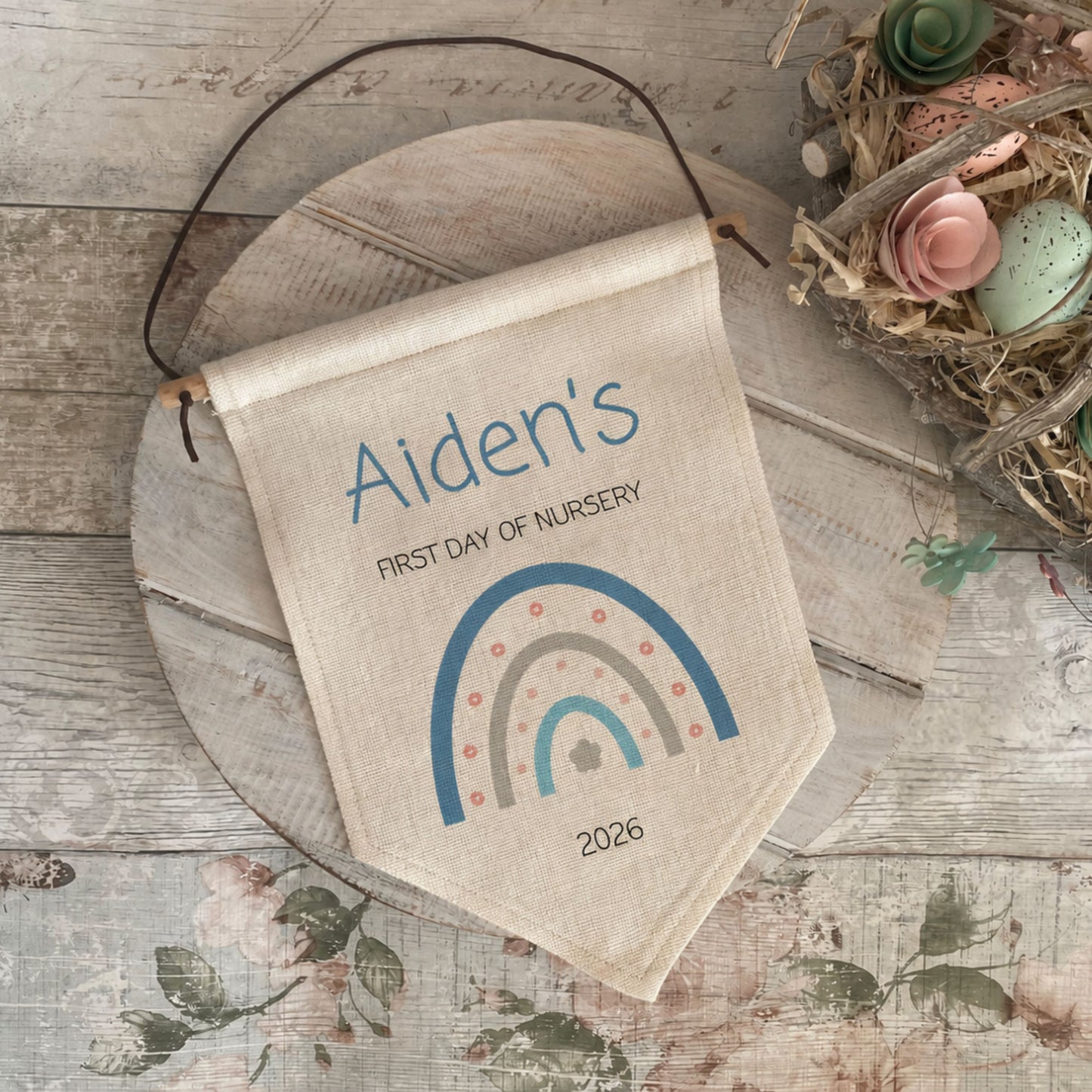 A personalized canvas nursery flag hanging on a rustic wooden board, featuring the text 'Aiden's FIRST DAY OF NURSERY' and a colorful rainbow design with the year 2026. The flag is styled in a nursery setting alongside a wooden tray and a decorative bird nest with pastel eggs.