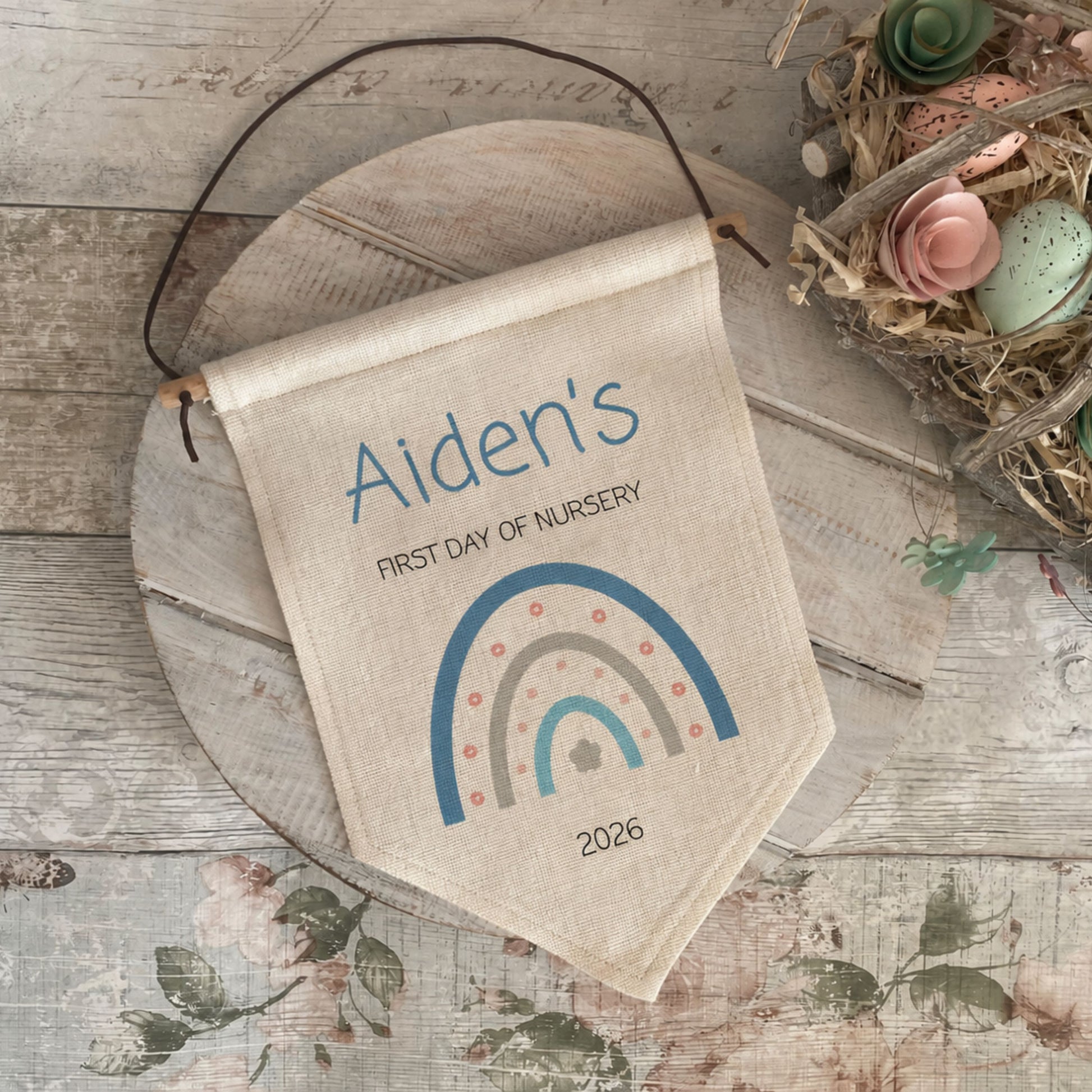A personalized canvas nursery flag hanging on a rustic wooden board, featuring the text 'Aiden's FIRST DAY OF NURSERY' and a colorful rainbow design with the year 2026. The flag is styled in a nursery setting alongside a wooden tray and a decorative bird nest with pastel eggs.