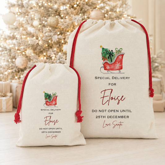 Two personalized Christmas gift bags in natural canvas with red drawstrings, featuring a red sleigh illustration and custom text, displayed in front of a blurred decorated Christmas tree.