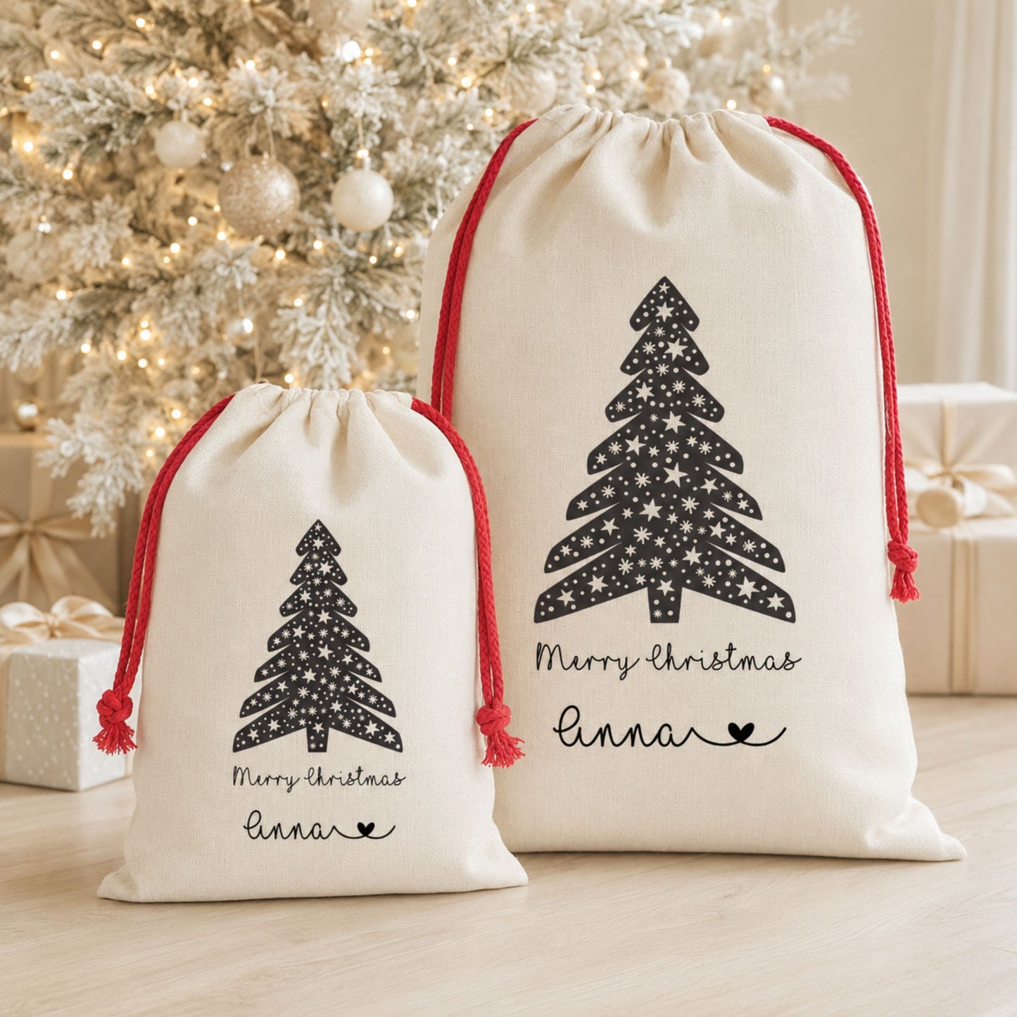 Two cream drawstring gift bags with red cords and black Christmas tree prints, displayed in front of a decorated Christmas tree and wrapped gifts on a light wood floor.
