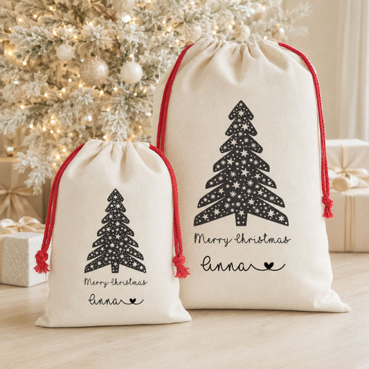 Two cream drawstring gift bags with red cords and black Christmas tree prints, displayed in front of a decorated Christmas tree and wrapped gifts on a light wood floor.