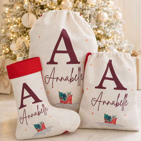 Three personalized Christmas gift bags and a matching stocking set in cream fabric, printed with a dark red monogram letter A, the name Annabelle, and a small red-and-blue gift illustration. The set is arranged in front of a softly blurred decorated Christmas tree.