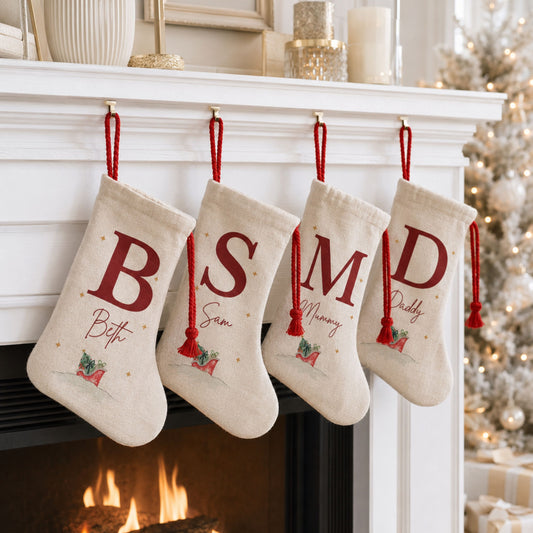 Four personalized Christmas stockings in cream fabric hang from a white fireplace mantel. Each stocking has a large red letter (B, S, M, D), a red tassel, and custom names embroidered below.