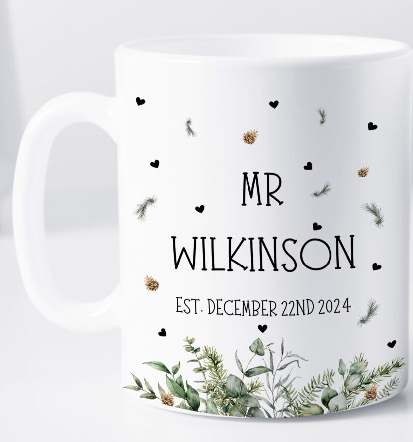 A white ceramic mug featuring the text "MR WILKINSON" and "EST. DECEMBER 22ND 2024" in black capital letters. The design includes scattered black hearts, green pine sprigs, and gold pinecones. At the base of the mug is an arrangement of illustrated winter greenery, including eucalyptus leaves and fir branches. The background is a clean, white surface.