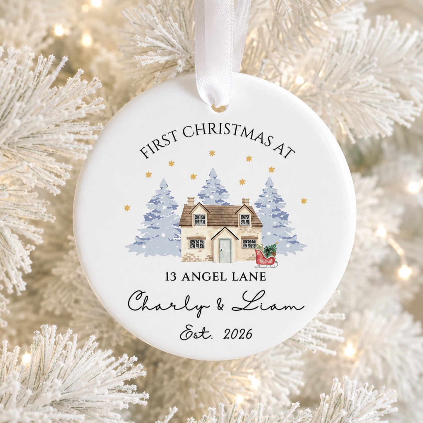 A round white personalized Christmas ornament hanging from a white ribbon, printed with the text 'FIRST CHRISTMAS AT 13 ANGEL LANE Charly & Liam Est. 2026' and a small illustration of a cozy house, trees, and stars.