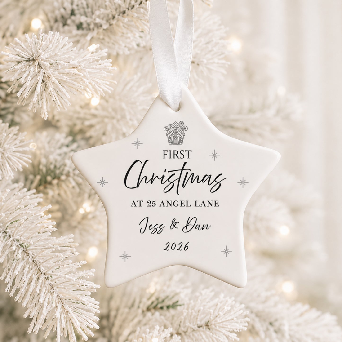 A white star-shaped Christmas ornament hanging from a white ribbon, printed with the personalized text 'FIRST Christmas AT 25 ANGEL LANE Jess & Dan 2026' and a small crest, shown in front of a softly blurred white frosted Christmas tree.