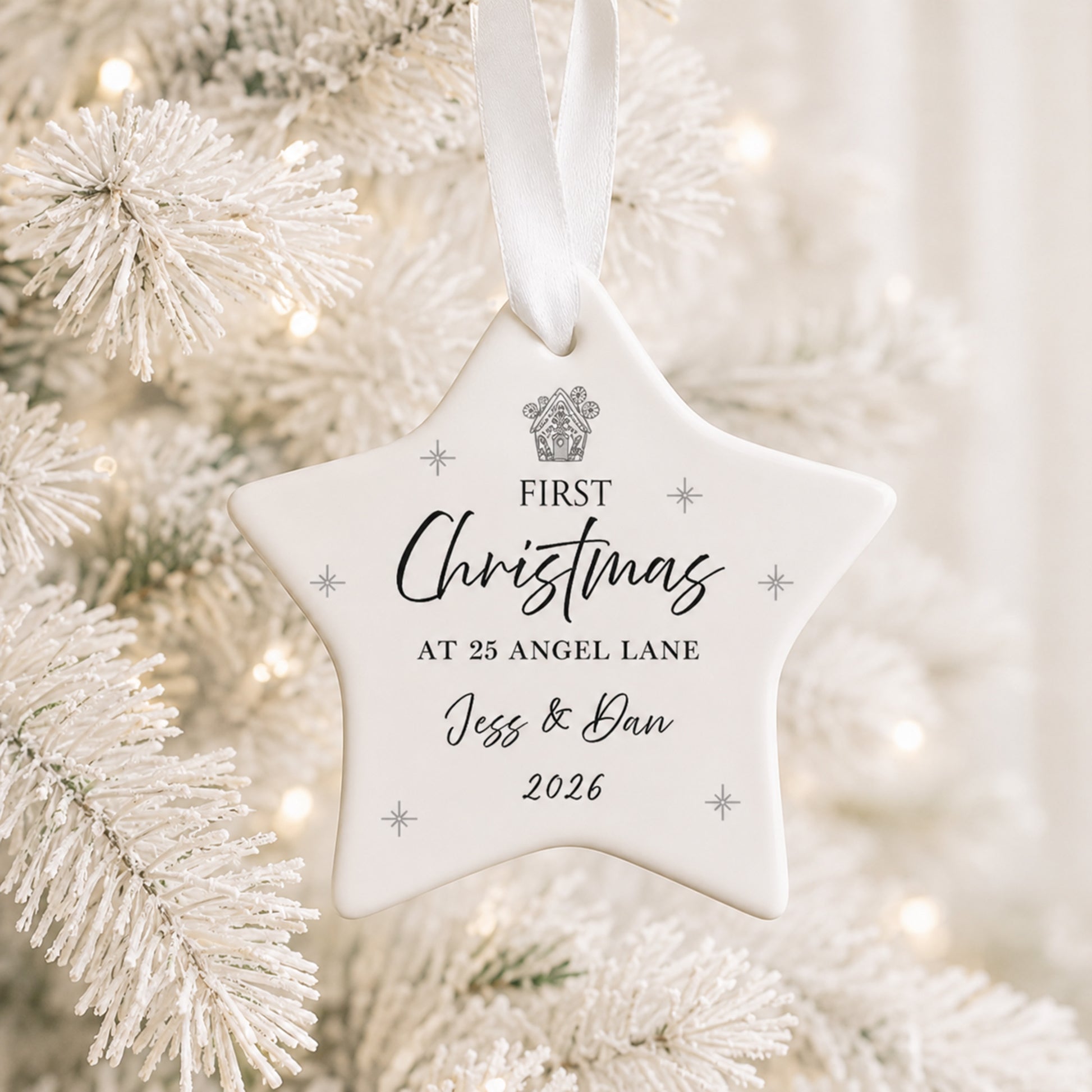 A white star-shaped Christmas ornament hanging from a white ribbon, printed with the personalized text 'FIRST Christmas AT 25 ANGEL LANE Jess & Dan 2026' and a small crest, shown in front of a softly blurred white frosted Christmas tree.