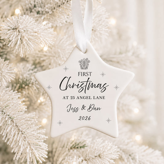 A white star-shaped Christmas ornament hanging from a white ribbon, printed with the personalized text 'FIRST Christmas AT 25 ANGEL LANE Jess & Dan 2026' and a small crest, shown in front of a softly blurred white frosted Christmas tree.