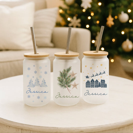 Three personalised mason jar tumblers with Christmas designs on a table in front of a decorated tree.