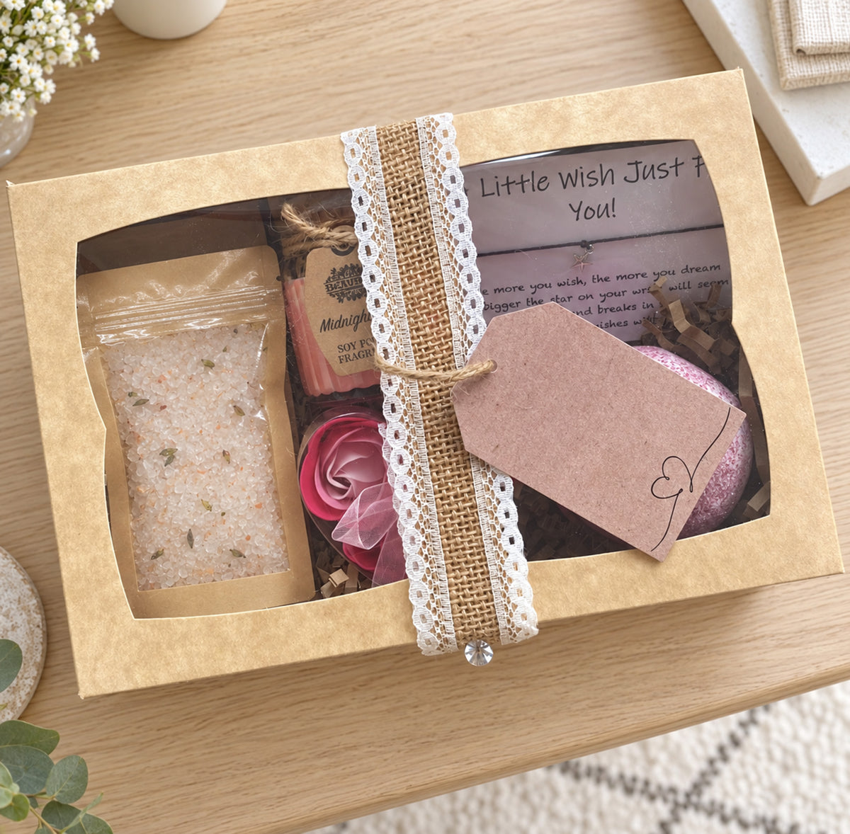 A curated bath gift box set displayed in a white box on a wooden table. The set includes a pink bath bomb, a pink rose-shaped soap, a soy rose candle, and a bag of bath salts, all nestled in brown paper shred with a handwritten 'A Birthday Wish' card.The box is tied with a lace and burlap ribbon and has a decorative tag.
