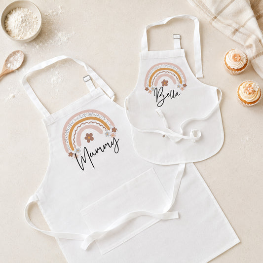 A matching apron set for two, featuring a larger apron labeled 'Mummy' and a smaller bib apron labeled 'Bella', both printed with a pastel rainbow and floral design, styled on a beige surface with baking props.