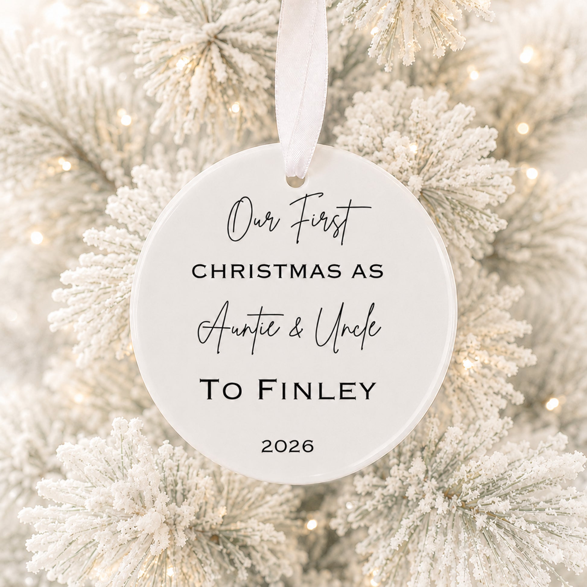 A round white personalized Christmas ornament hanging from a white ribbon, printed with the text 'Our First CHRISTMAS AS Auntie & Uncle TO Finley 2026' in black script and serif lettering, set against a blurred background of a frosted white Christmas tree with warm string lights.