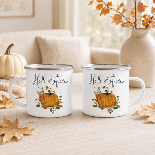 Two white enamel-style mugs with silver rims sit on a light wood table, each printed with the words “Hello Autumn” above an orange pumpkin and fall leaves. The scene is styled with autumn leaves, a small pumpkin, a beige sofa, and a vase in the background.