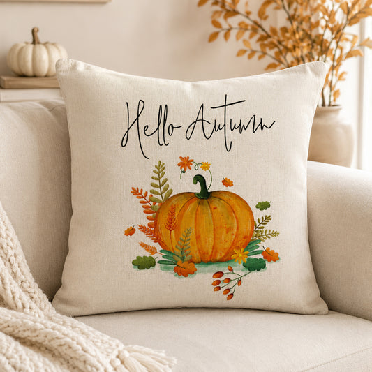 A square cream throw pillow with a watercolor autumn design featuring a pumpkin, leaves, and the text 'Hello Autumn' in black script, styled on a beige sofa with a knit blanket.