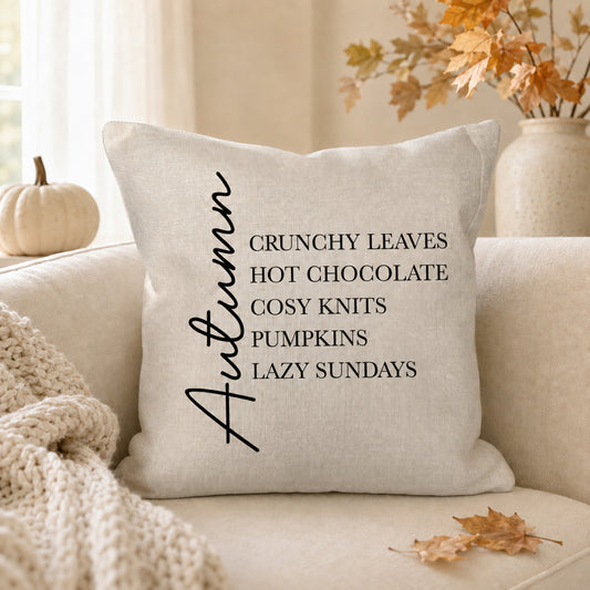 A square beige throw pillow with black text reading 'Autumn CRUNCHY LEAVES HOT CHOCOLATE COSY KNITS PUMPKINS LAZY SUNDAYS' sits on a cream sofa in a cozy autumn-themed room with a knit blanket, a small pumpkin, and fall leaves.