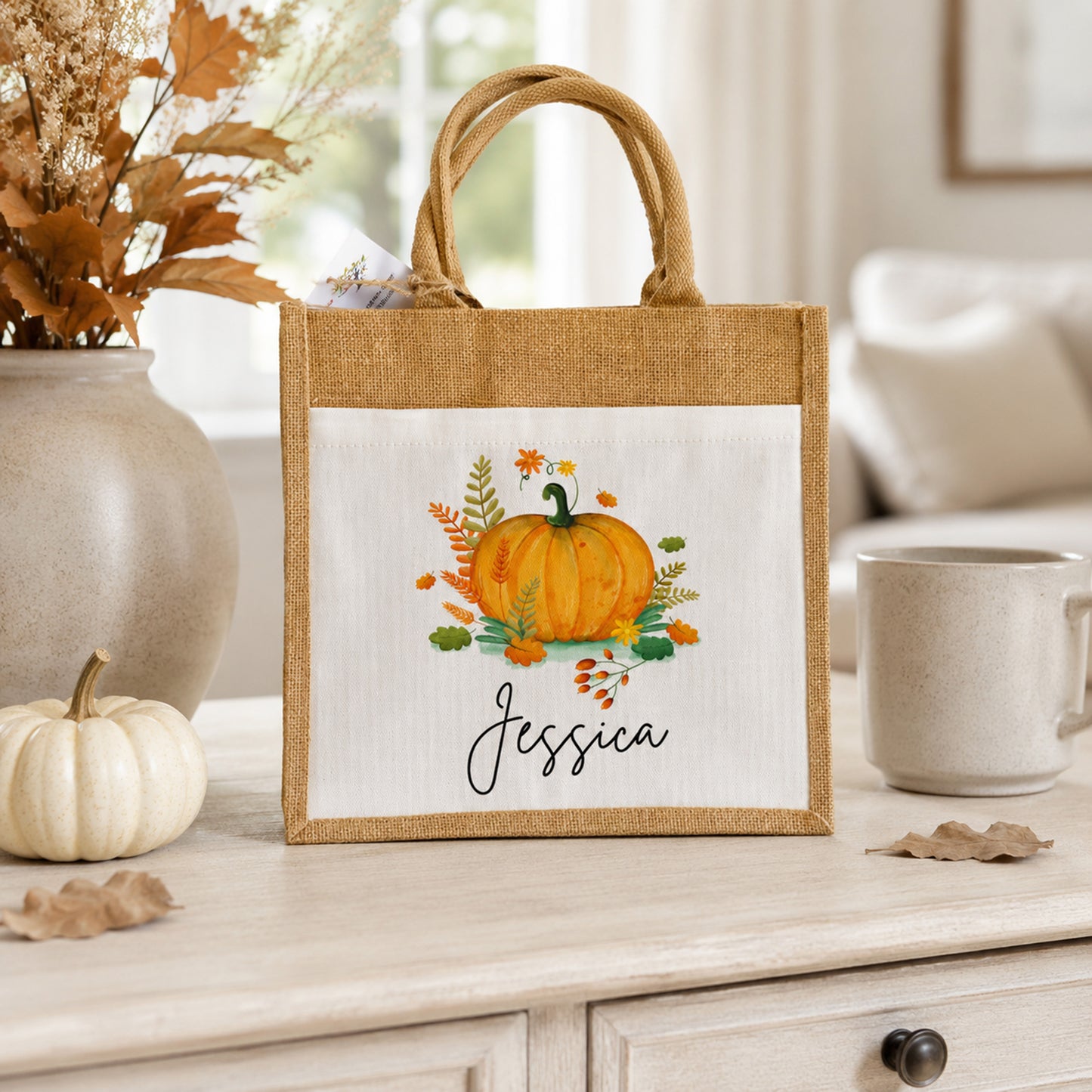A personalized burlap tote bag with a white front panel featuring a watercolor pumpkin illustration and the name 'Jessica' in script, styled on a light wood table with autumn decor.