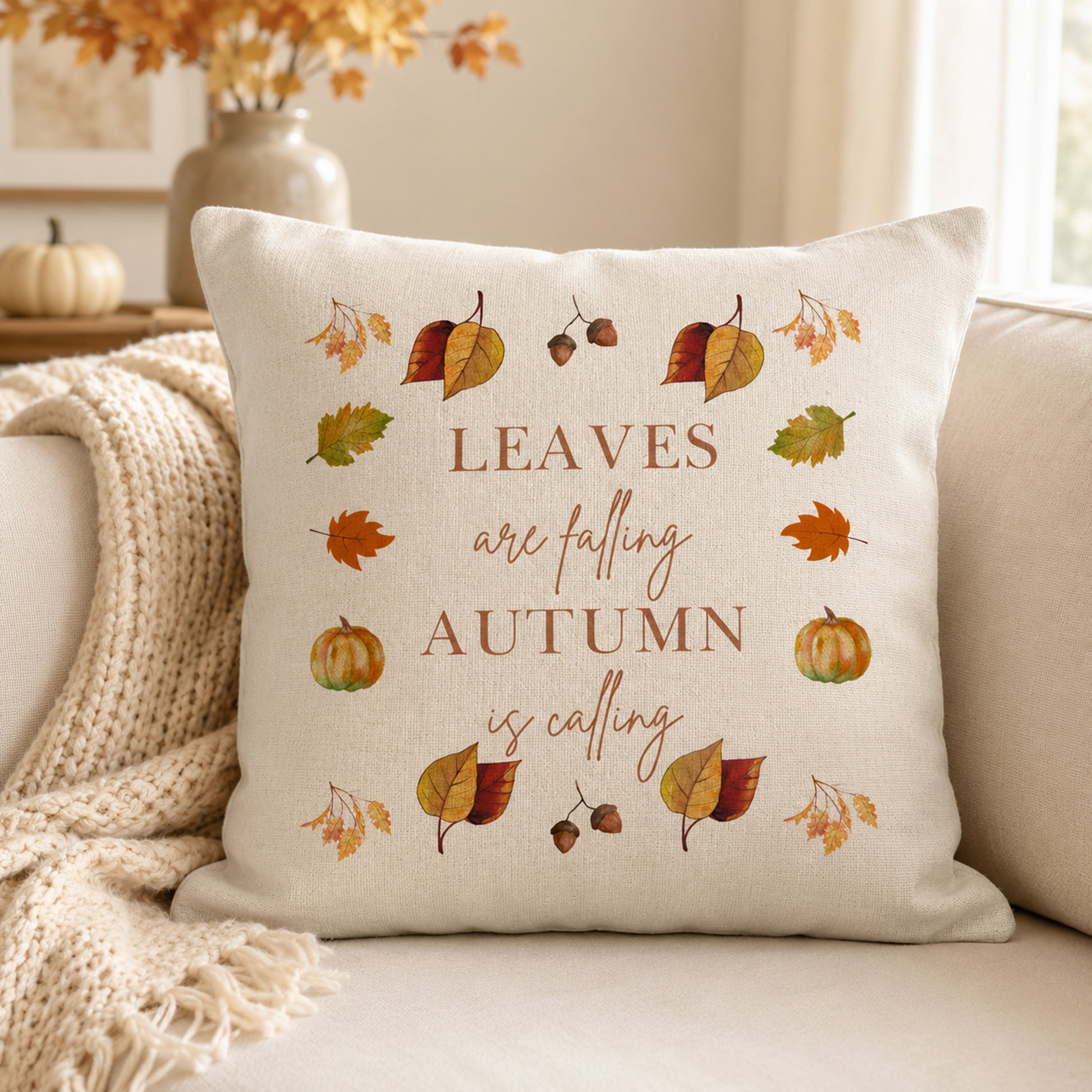 A square cream throw pillow with an autumn-themed design featuring the phrase 'LEAVES are falling AUTUMN is calling' in brown lettering, surrounded by illustrated leaves, acorns, and pumpkins. The pillow is styled on a beige couch with a cream knit throw blanket, with a blurred background showing a vase of fall leaves and a small pumpkin.