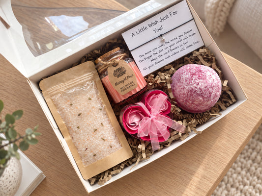 A curated bath gift box set displayed in a white box on a wooden table. The set includes a pink bath bomb, a pink rose-shaped soap, a soy rose candle, and a bag of bath salts, all nestled in brown paper shred with a handwritten 'A Little Wish' card.The box is tied with a lace and burlap ribbon and has a decorative tag.