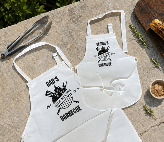 Two white aprons laid flat on a stone surface, each printed with a black barbecue-themed graphic and text. One apron reads "DAD'S BARBECUE" and the other reads "GEORGE'S BARBECUE.