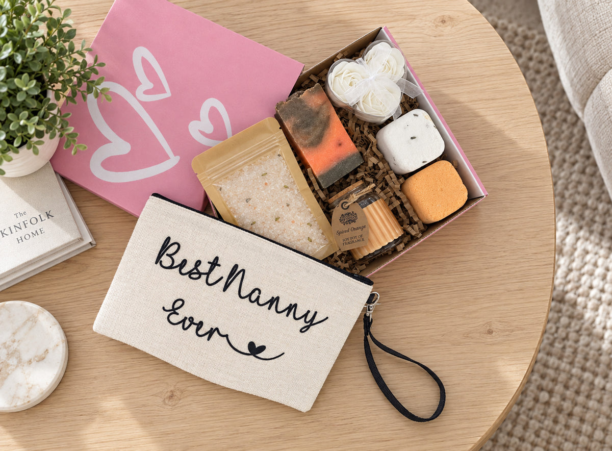 A curated gift set arranged on a round wooden table, featuring a cream canvas pouch with 'Best Nanny Ever' embroidered in black script, a pink gift box with white heart outlines, and an open box containing bath salts, soap bars, and a candle.