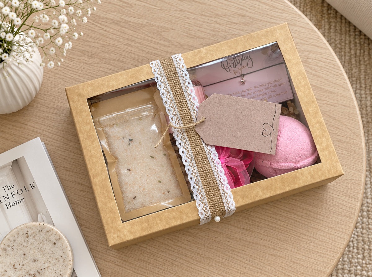 A square kraft gift box with a clear window, tied with burlap ribbon and lace trim, containing bath or body items including bath salts, a pink rbath bomb, and a kraft gift tag. The box sits on a light wood table with decorative props nearby.