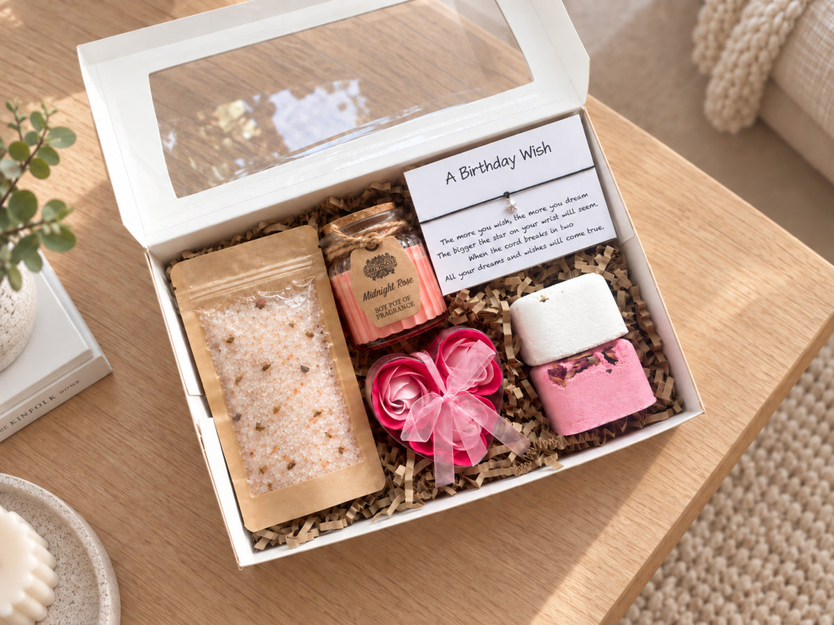 A curated gift box containing a bath salt packet, a candle, a pink flower-shaped soap, and a handwritten birthday wish bracelet, arranged in shredded paper inside an open white box on a wooden table.