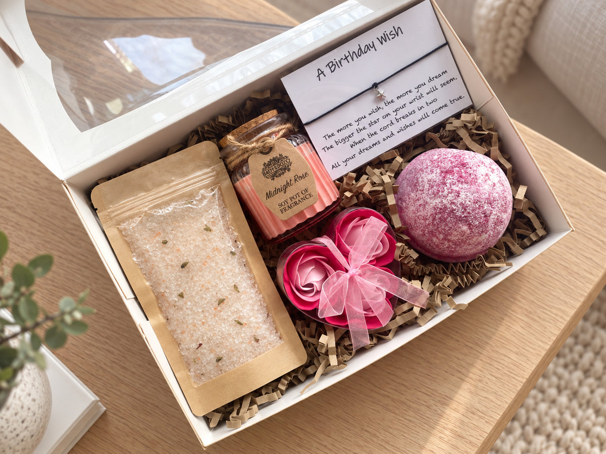 A curated bath gift box set displayed in a white box on a wooden table. The set includes a pink bath bomb, a pink rose-shaped soap, a soy rose candle, and a bag of bath salts, all nestled in brown paper shred with a handwritten 'A Birthday Wish' card.