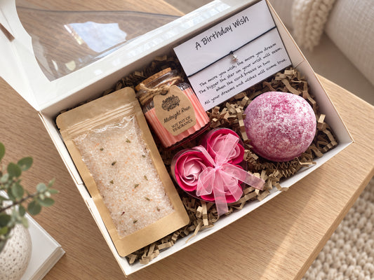 A curated bath gift box set displayed in a white box on a wooden table. The set includes a pink bath bomb, a pink rose-shaped soap, a soy rose candle, and a bag of bath salts, all nestled in brown paper shred with a handwritten 'A Birthday Wish' card.