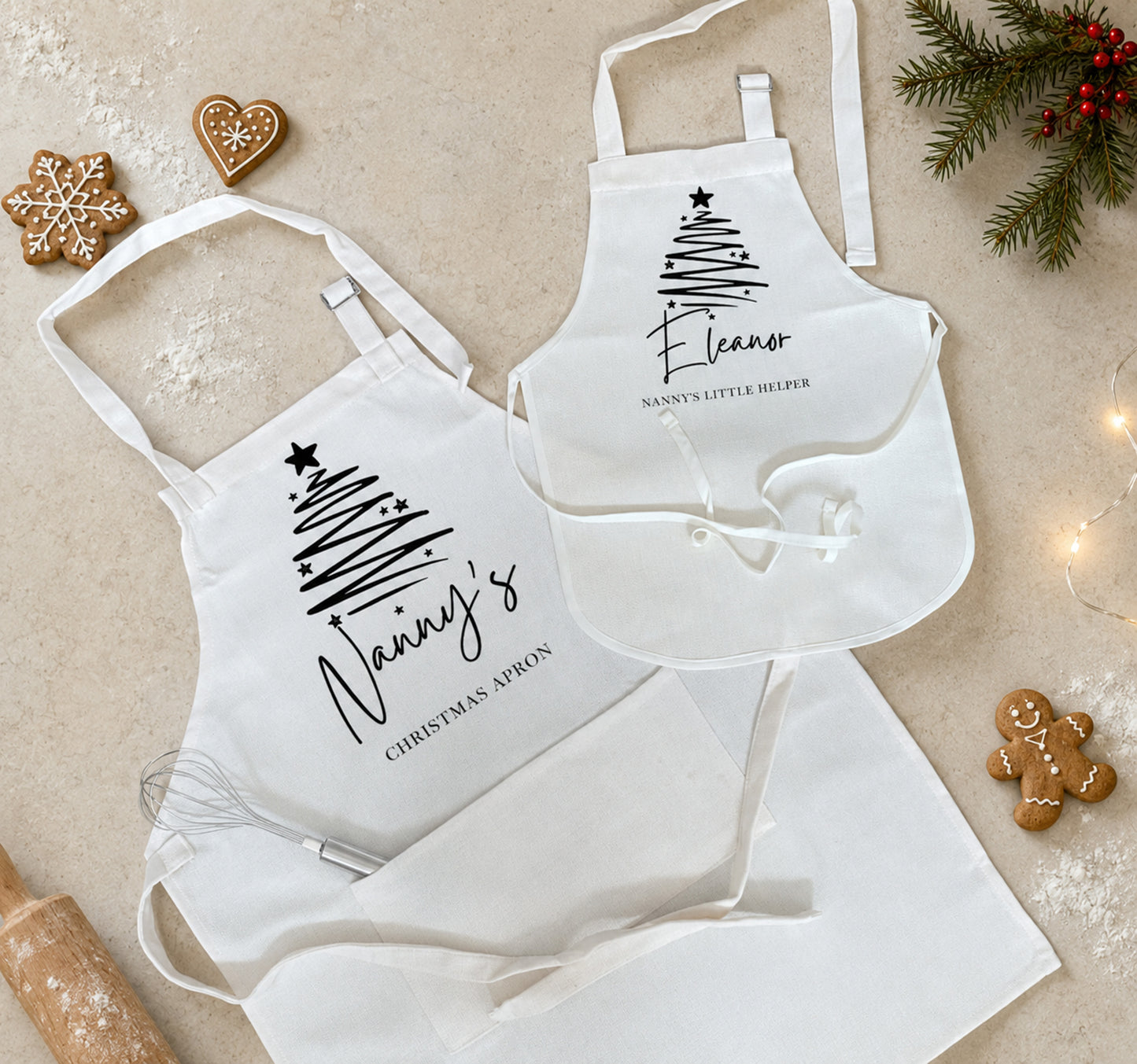 Three white aprons arranged on a light beige surface with holiday baking props, including gingerbread cookies, evergreen branches, and string lights. Two aprons feature black printed Christmas tree graphics and names, while one larger apron is plain white.