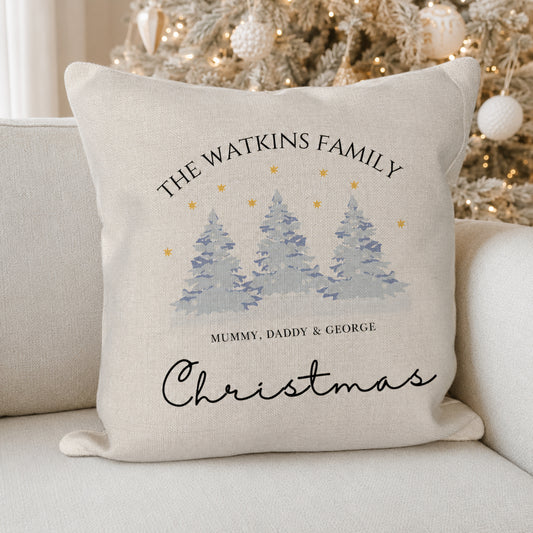 A square cream-colored linen throw pillow with a personalized Christmas design, resting on a light beige sofa with a blurred Christmas tree in the background. The pillow features the text 'THE WATKINS FAMILY' and 'MUMMY, DADDY & GEORGE' above an illustration of three snowy pine trees with gold stars, and 'Christmas' written in black cursive at the bottom.