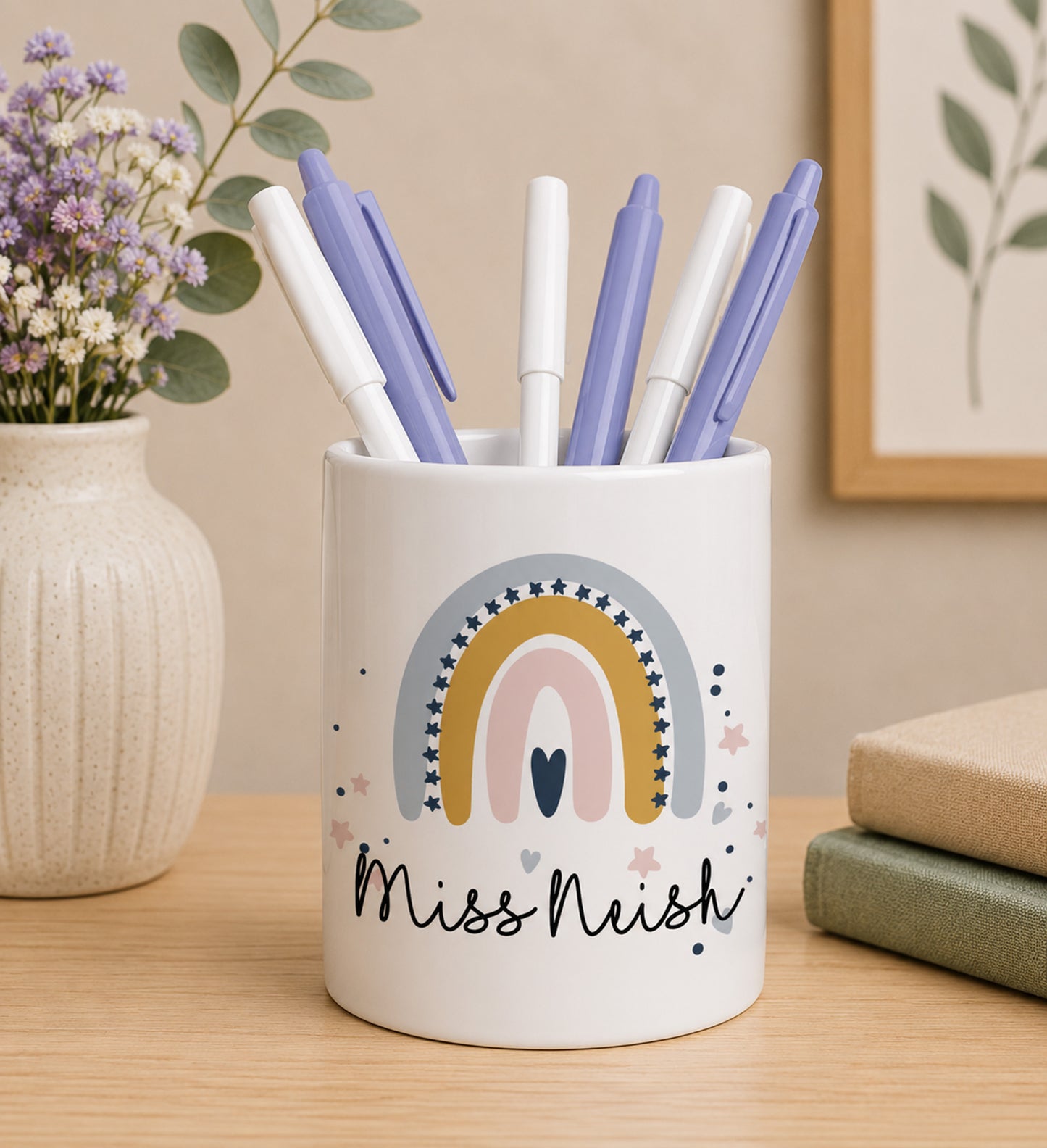White teacher pencil pot with colourful rainbow design and custom name 'Miss Nishi' text, filled with pens on a wooden surface.