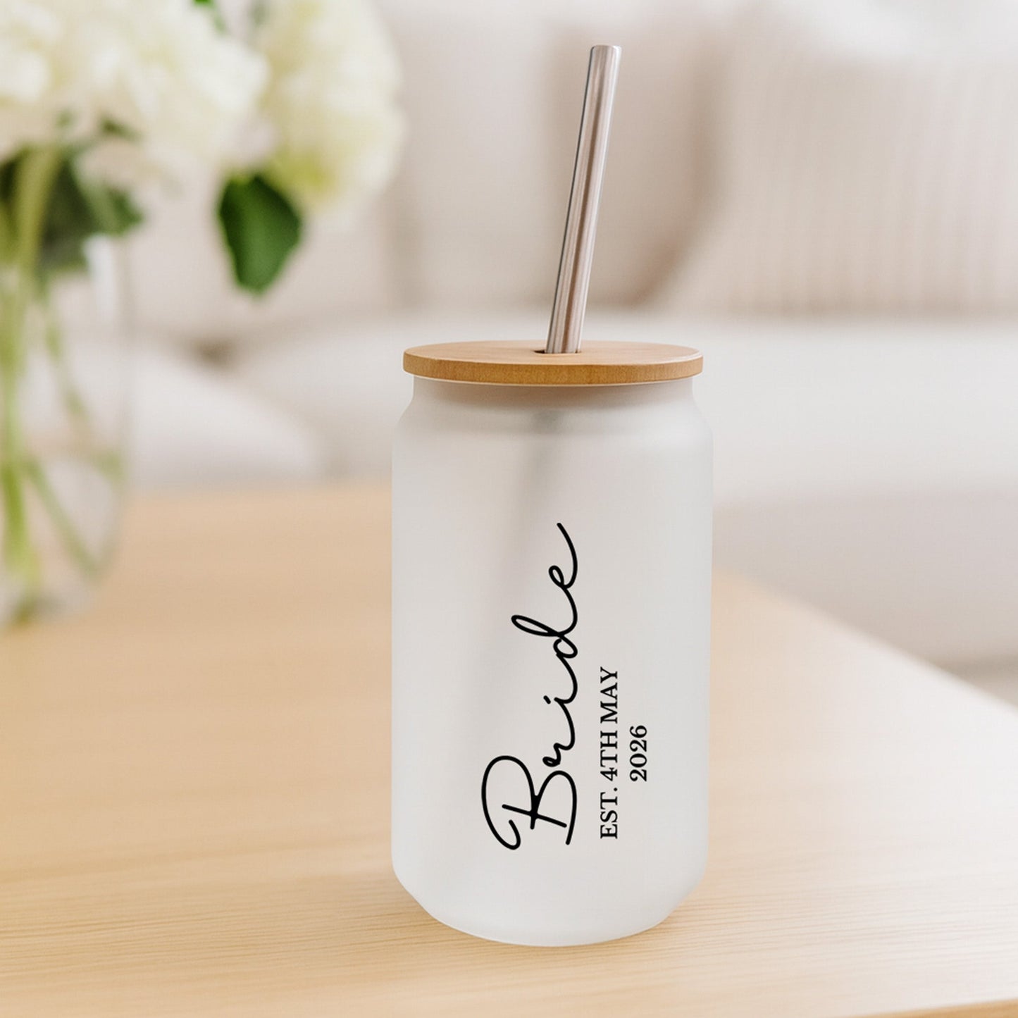 Glass tumbler with 'Bride' text and wooden lid on a light surface with flowers in the background