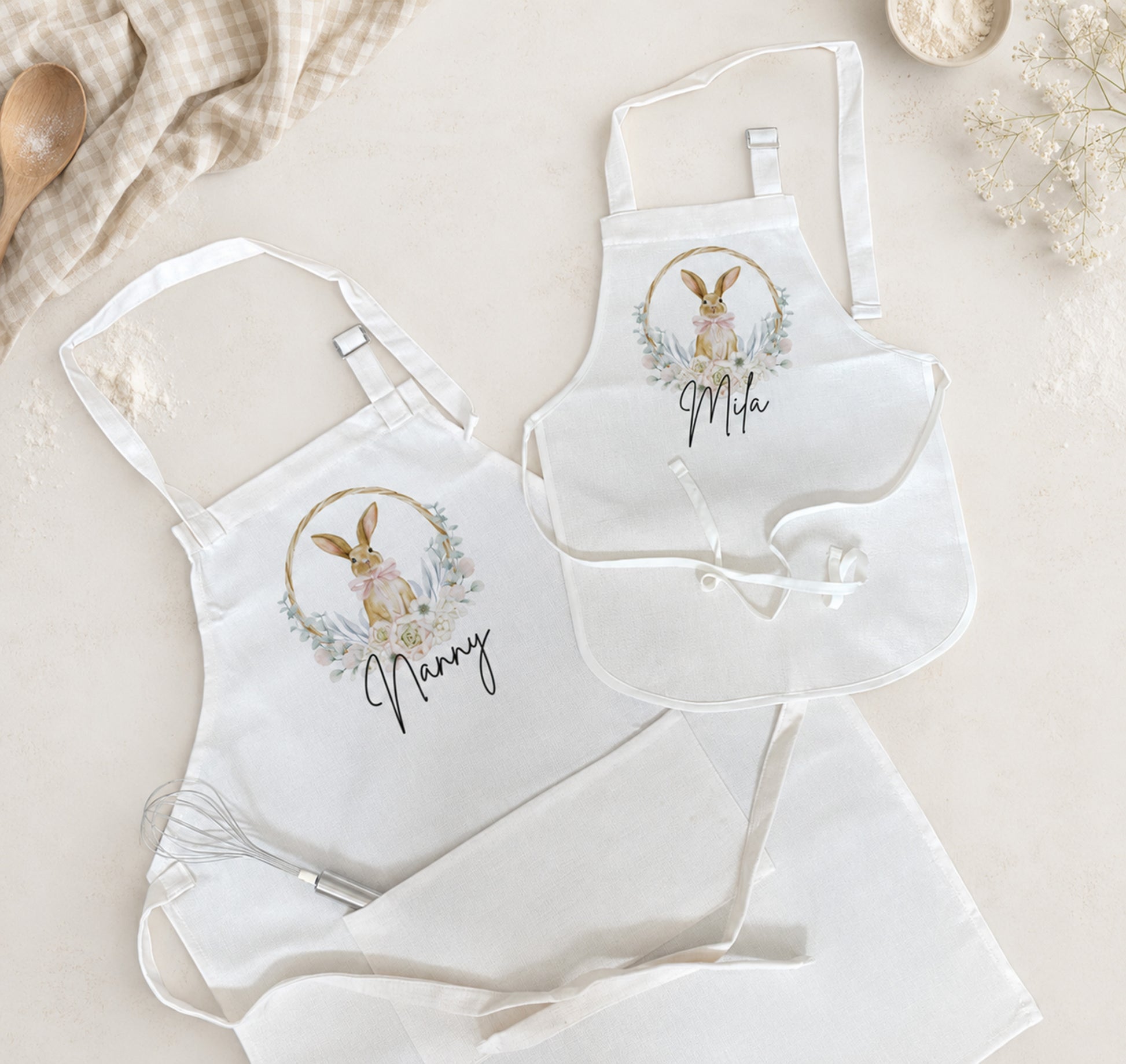 A coordinated set of white aprons laid flat on a light beige surface, including two adult aprons and one smaller apron. Each apron has a watercolor-style bunny wreath graphic and the name printed below in script.