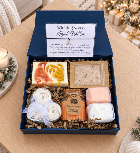 A Christmas-themed gift box containing handmade soaps, a bath salt packet, and a handwritten note, displayed on a wooden table with holiday decorations in the background.