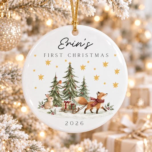 A personalized round ceramic Christmas ornament hanging from a gold string, featuring the text 'Erin's FIRST CHRISTMAS 2026' and an illustration of a fox pulling a sleigh with gift boxes near decorated trees. The ornament is shown hanging in a softly blurred festive tree setting with warm lights and wrapped gifts.