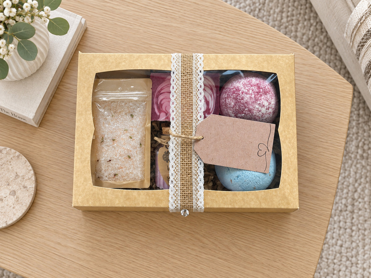 A gift box containing bath products, including a clear bag of bath salts and colorful bath bombs, tied with a lace ribbon and a kraft paper tag, displayed on a wooden table.