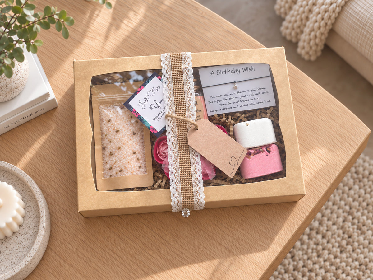 A curated gift box containing a bath salt packet, a candle, a pink flower-shaped soap, and a handwritten birthday wish bracelet, arranged in shredded paper inside an open white box on a wooden table.