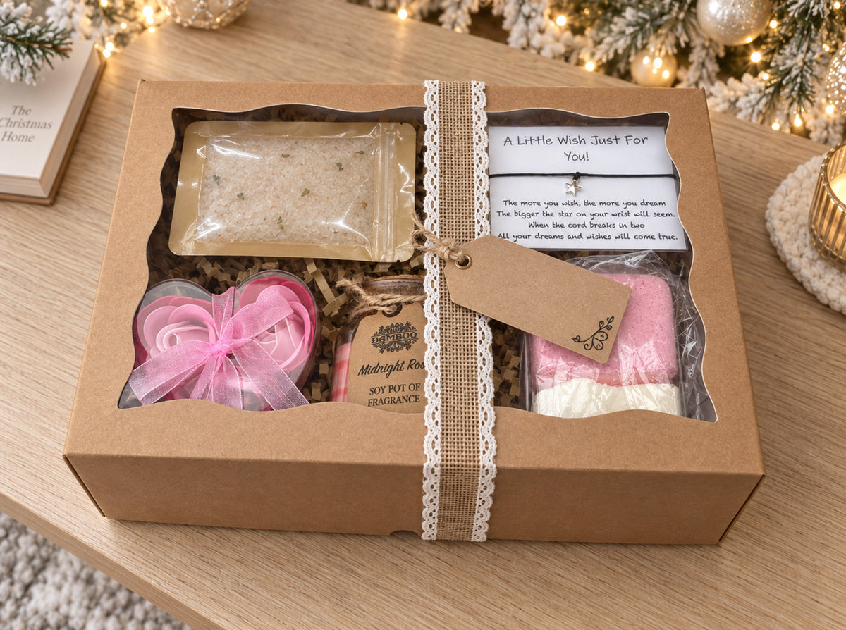 A Christmas-themed gift set in a brown kraft box with a lace ribbon, containing shower steamers, a pink heart-shaped soap flowers, a small candle and bath salts, displayed on a wooden table with warm holiday lights in the background.
