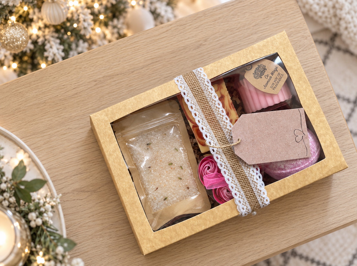 A gift box containing a clear pouch of bath salts, pink rolled bath soap, and a pink bath bomb in a kraft paper box with a lace ribbon and kraft tag, styled on a wooden table with holiday lights in the background.