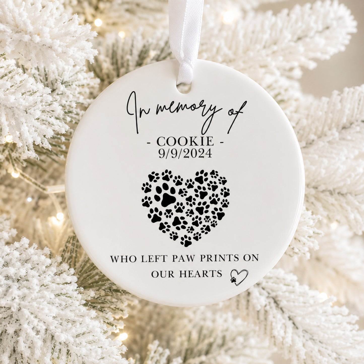 A round white memorial Christmas ornament hanging on a white-frosted tree branch, printed with the text 'In memory of - COOKIE - 9/9/2024' and a heart made of black paw prints, with additional text 'WHO LEFT PAW PRINTS ON OUR HEARTS'.
