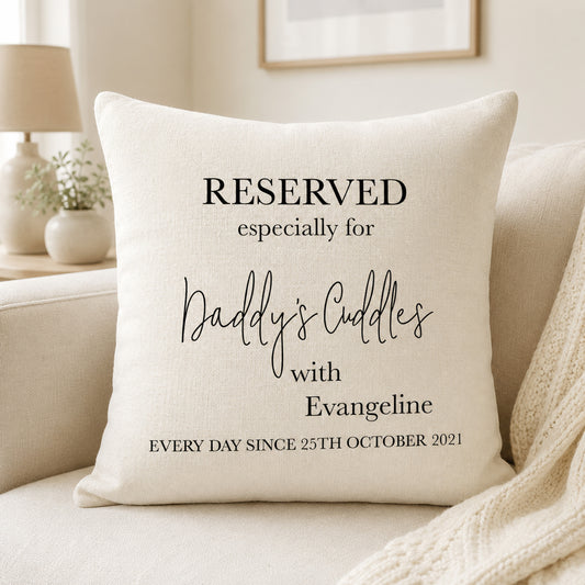 A square cream throw pillow with black printed text reading 'RESERVED especially for Daddy's Cuddles with Evangeline EVERY DAY SINCE 25TH OCTOBER 2021', styled on a beige sofa in a softly lit living room.