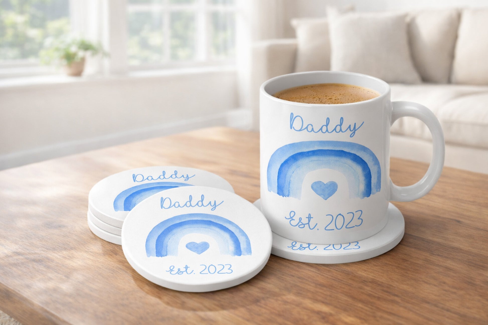 White mug and coasters with blue rainbow design and 'Daddy est 2023' text on a wooden table.